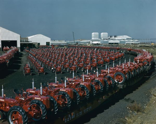 Elevated view of a large group of Farmall C tractors parked outdoors in rows at Louisville works ready to be shipped. Near the center is a man with his knee up on a front wheel of a tractor writing on a clipboard. In the foreground tractors are mounted on railroad cars on railroad tracks.