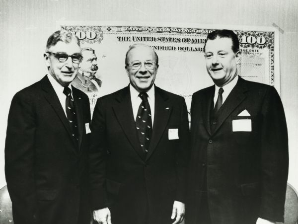 Brooks McCormick standing with two unidentified men. Behind them is an enlarged reproduction of a United States Savings Bond.