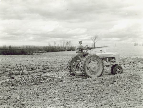 Right side view of a man driving a Farmall H tractor pulling a spring tooth harrow. George Cabak farm of 200 acres.