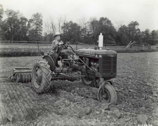 Three-quarter view from front left of a 10-year-old boy, Wilmer Nissley, driving a Farmall B tractor with a disk harrow attachment. He is driving his father's new Farmall B on a 101-acre farm in Lancaster County.