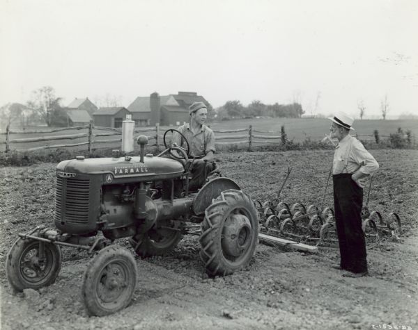 Amos Shelby, Jr. sitting on a Farmall A tractor on his father's 80-acre farm near Manheim. He is talking with J.B. Hostetter, of Mount Joy, International Harvester Dealer, who is standing on the right. Farm buildings are in the background.