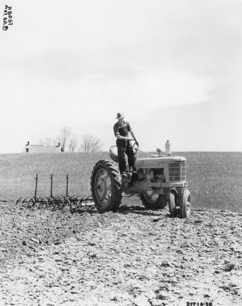 J.T. Tully standing and driving a Farmall H tractor and 3 section spring-tooth harrow in a field. Peterborough, Ontario, Canada.