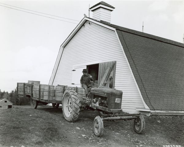 Alden G. Bagley driving a Farmall M pulling a wagon loaded with barrels of seed potatoes. Behind him is his 6,000 barrel storage house.