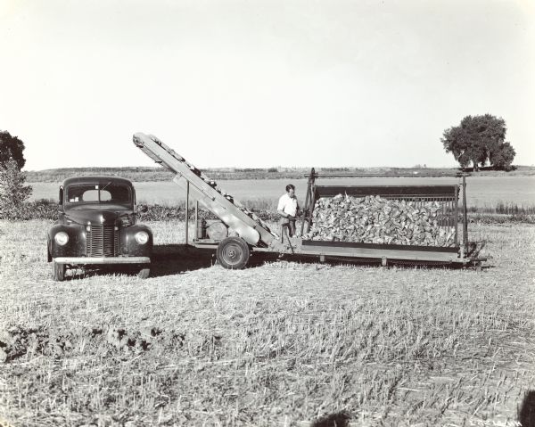 View of a man raising platform of stationary loader by ratchet. This moves the beets to conveyor operated by small 3-horsepower Model LB engine. An International truck is parked on the left under the conveyor.