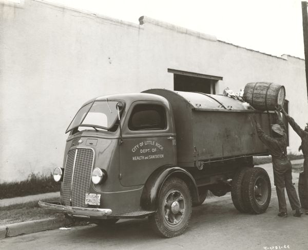 Three-quarter view from front left of a D-300-H truck used for garbage collection. Hercules body hydraulic hoist. Rounded sliding covers. Waterproof tailgates. Two men are dumping a barrel into the back of the truck parked near the side of a building. Sign painted on driver's side door reads: "City of Little Rock."