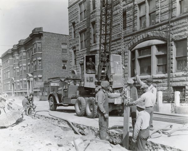 View from street of men working on a Works Progress Administration project. A group of men are standing and holding a beam upright in a trench, with a chain attached to it suspended from a crane parked behind. One man operates the crane, and another man sits in the driver's seat of truck, a six-wheeler, carrying the crane. Other men are working with a hose on the left. A sign on the back of the truck reads, in part: "District 3 Chicago, Penalty for Theft or Destruction, Fine Not to Exceed 10,000 on Imprisonment Not to Exceed 10 Yrs."