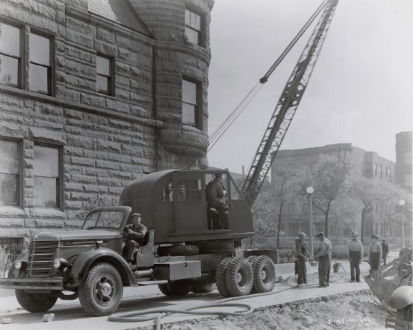 View across street towards men working on a Works Progress Administration project. Three men are standing and holding a board with a chain attached to it near a crane parked in the street on the left. One man is operating the crane, and another man is sitting in the driver's seat of truck, a six-wheeler, carrying the crane.