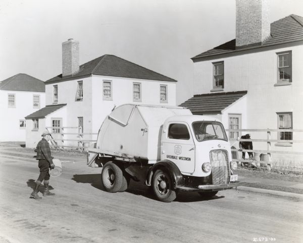 View of man carrying a metal trash bin towards a dump truck. A man sits in the driver's seat. Another man stands near metal trash bins behind a fence along a row of houses. A sign on the passenger side door reads: "U.S. Department of Agriculture. Farm Security Administration. Greendale Wisconsin."