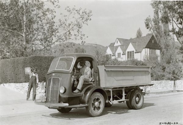 Two men, one in the driver's seat of the D-300 M.T. truck, and the other carrying a metal trash bin, on a residential street. The truck is used by the Refuse Disposal Department for pickup of garbage in divided streets or on hilly and winding roads. Pasadena purchased its first International in 1935 — now have 30 in operation. A large house and hills are in the background.