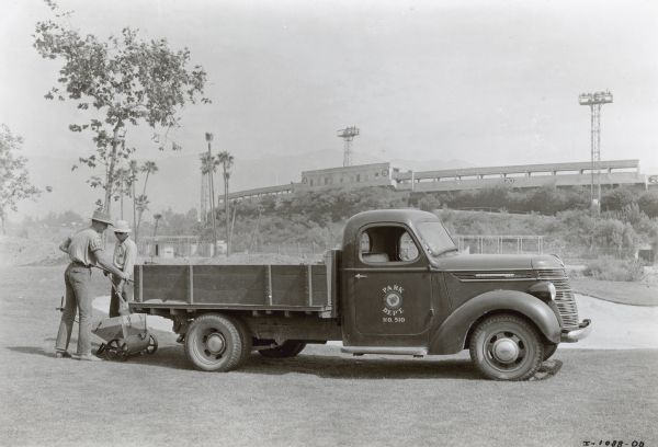 Two men are standing near the back of a Model D-15 owned by the City of Pasadena parked on the municipal golf course. The truck is used by the Park Department. In the background is the Rose Bowl Stadium, with mountains in the far background.