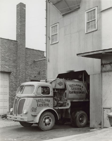 A man sitting in the driver's seat of a DR-500 with 94 inch wheelbase. Blair Knox 2 1/2 cubic yard truck mixer. FF-42-C direct in fifth transmission. Painted sign on truck reads: "Ireland and Lester, Ready Mixed Concrete."