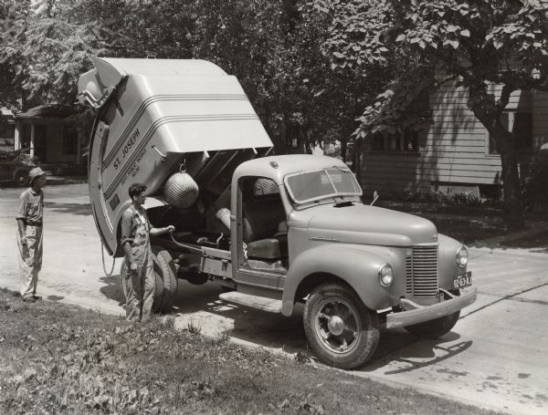 View across lawn towards two men standing near a K-5 truck used by the Department of Public Works, St. Joseph. The doors of the cab are removed. One man is operating the hoist so that the garbage slides down from front to rear.