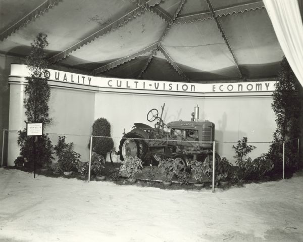 View of display inside a tent at the Minnesota State Fair of a McCormick-Deering Farmall A tractor set up in front of a wall with a sign above it reading: "Quality Culti-Vision Economy."
