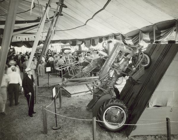 Slightly elevated view of Farmall B tractor on display at the Missouri State Fair. The tractor is leaning at an angle against a ramp. A crowd of people are in the open sided tent looking at the displays, which also include trucks and agricultural machinery. 