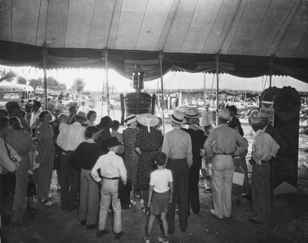 Slightly elevated view of crowd inside an open sided tent gathered in front of "Harvey Harvester." There is a Farmall tractor parked outside of the tent on the right.