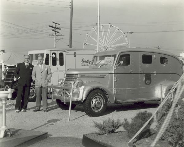 Mr. Collis Dell and Mr. Allen of the Brink's Company stand outdoors with the Brink's Express Money Truck. The Brink's streamlined money wagon created so much attention that it was necessary to chain if off to keep the people from climbing on the fenders, hood, and roof. In the left foreground is the water fountain which was part of the front entrance of the truck display.