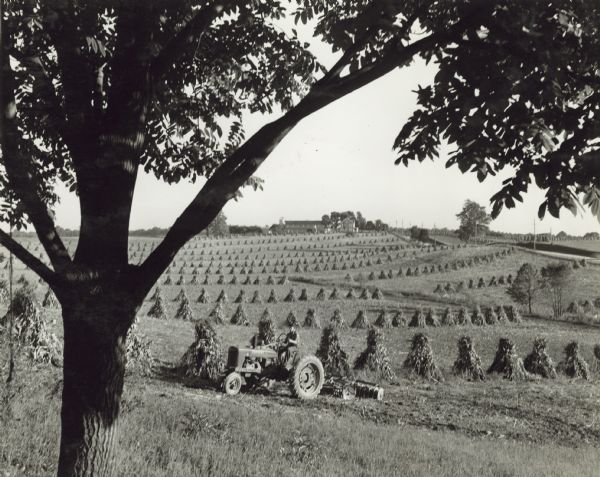 Left side view of a man driving a Farmall H tractor with disk harrow in a field. Shocks of corn are in long rows in the field. Farm buildings are in the far background. James Clark Farm.