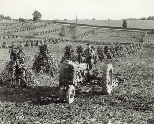 Three-quarter view from front left of a man driving a Farmall H tractor with disk harrow in a field. Shocks of corn are in rows in the field. James Clark Farm.