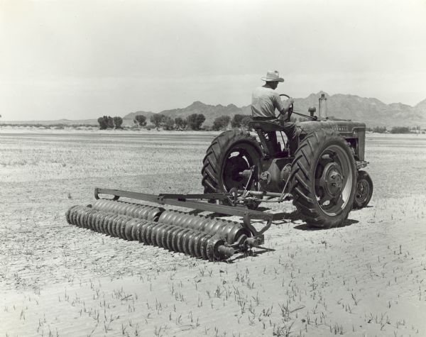 Three-quarter view from right rear of a man driving a Farmall M tractor pulling a pulverizer covering and packing residual alfalfa land. Arnold Travis.