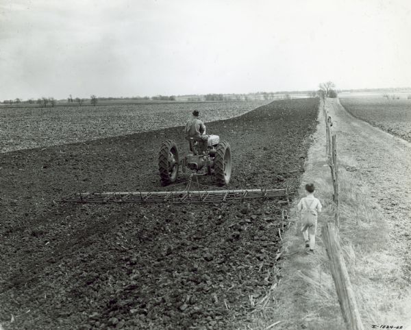 View down field towards a 15 foot peg tooth harrow being pulled by a man driving a Farmall H tractor. A young boy runs along a fence line on the right just behind the harrow. Owned by Glen Hernes.