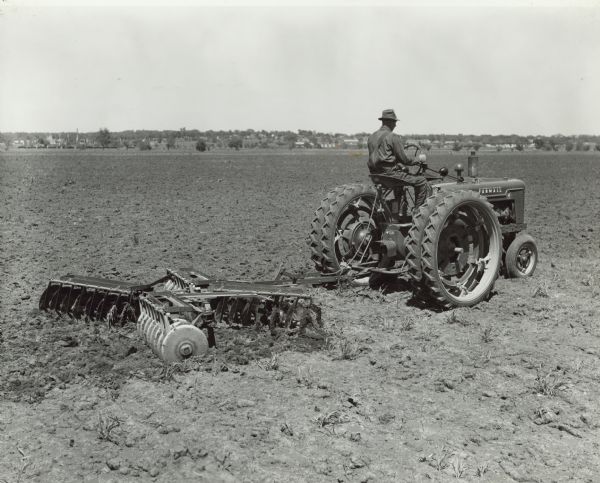 Three-quarter view from right rear of a man driving a Farmall H tractor with double wheels, pulling an 8-foot tandem disk. Mr. Joe Eichoff is preparing frozen-out wheat ground for replanting of corn.