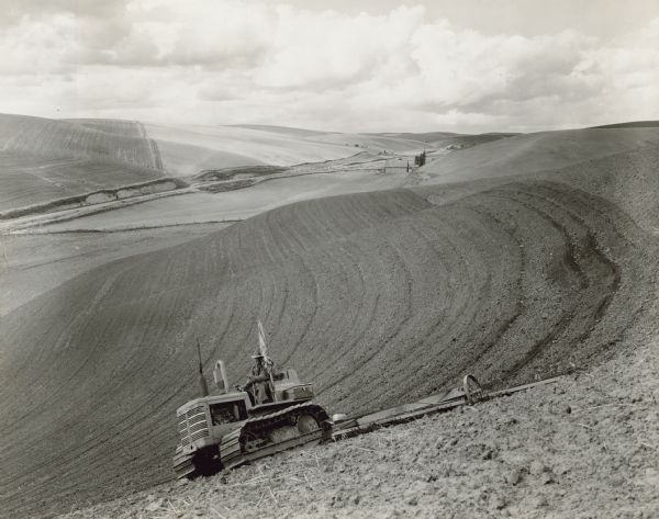 View down hill towards a man driving a TD-6 tractor with peg-tooth harrow on steep hills in wheat land. 