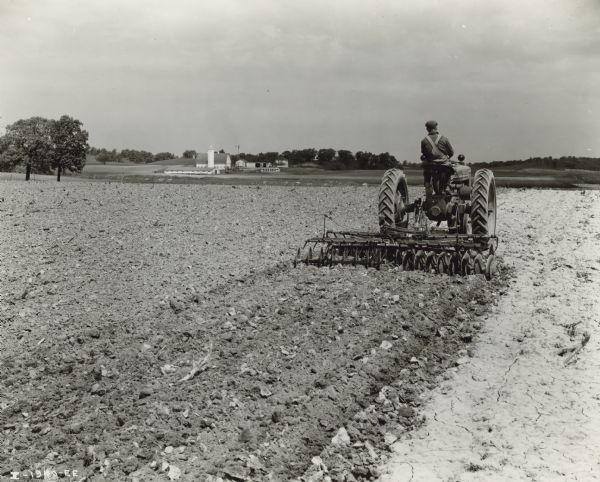 Rear view of a man, Harold Mills, driving a Farmall H tractor with a 9 foot McCormick-Deering disk harrow in a field. William Tobin farm. Farm buildings are in the far background on the left.