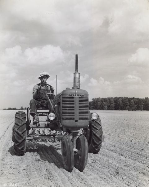 Front view of Harold Wyant driving a Model B Farmall tractor in a field. He is loosening up the soil in this field of young corn with a rotary hoe.