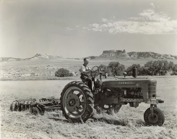 Left side view of Wayne Pattison driving a Farmall H tractor pulling a McCormick-Deering disc 7 in a field.