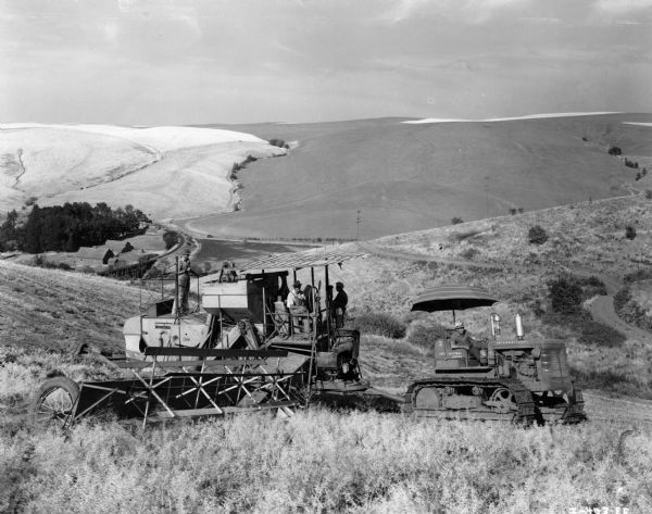 View down hill towards a group of men with a TD-14 and 51 combine owned by Eaton Sisters. They are harvesting mustard. Below them in the valley are farm buildings near railroad tracks going over a bridge.