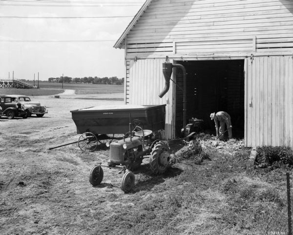 Elevated view looking down at a man using a Farmall Cub tractor to belt-drive a corn sheller. The man is inside the open doorway of a farm building bent over while shoveling the corn into the sheller. The Farmall cub is just outside the door. A McCormick-Deering wagon is on the left of the open doorway, receiving the shelled corn from a chute attached to the sheller. Automobiles are parked in the background on the left, and in the far distance is are stands next to a field. 