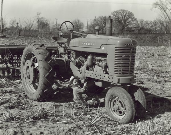 Young boy kneeling in a field and holding a hammer up near a Farmall M tractor pulling a grain drill. The boy is 3-years-old and is the son of the caretaker, Basil Allen. Major Albert A. Haan farm.