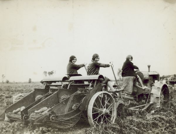 Three-quarter view from right rear of three women posing on a tractor pulling a potato digger outdoors in a potato field on the farm of L. Wilson. One woman is sitting in the tractor seat, and the other two women sit on a potato harvester, each holding a long handle. 