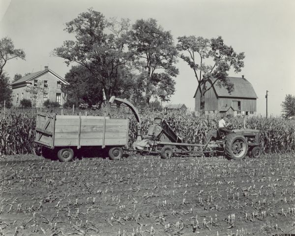View across harvested field towards a man using a wagon and an ensilage harvester drawn by a Farmall M tractor on 132 acre farm of A.W. Reisseeum. In the background are a farmhouse, automobiles, and farm buildings.