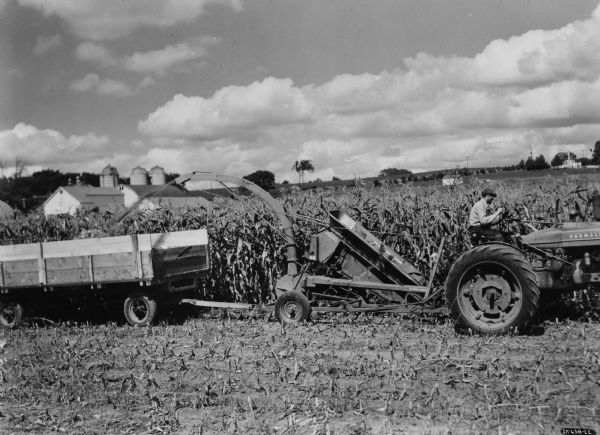 View across harvested cornfield towards a man using a Farmall M tractor to pull a No. 2 ensilage harvester and wagon and box on the Bowman Dairy Farm. Farm buildings and silos are in the far background.