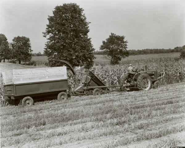 View across section of harvested field towards a man using a Farmall M tractor to pull a No. 2 ensilage harvester and McCormick-Deering wagon to cut corn on A. Wagner's farm.