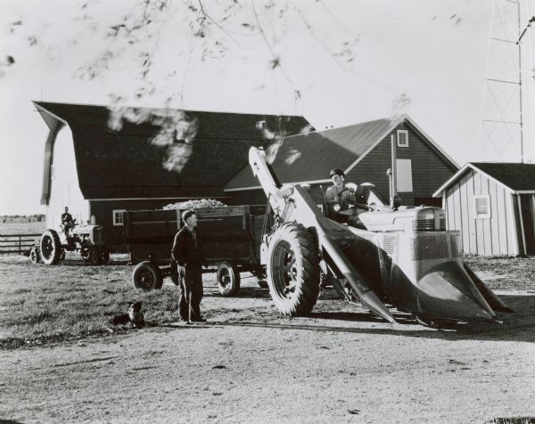 View of farmyard and farm buildings, with Clarence Schoger on a Farmall MD tractor with IHC corn picker and a wagon filled with ears of corn. Carl Schoger stands to the left of the tractor, and a dog is sitting on the ground nearby. In the background sitting on another tractor is Harry Schoger. 