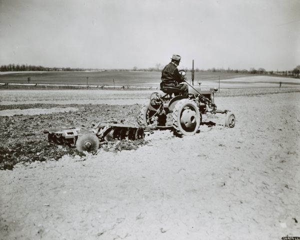 View across field towards a man using an experimental Farmall Cub pulling a four-foot tandem disk-harrow on a farm.