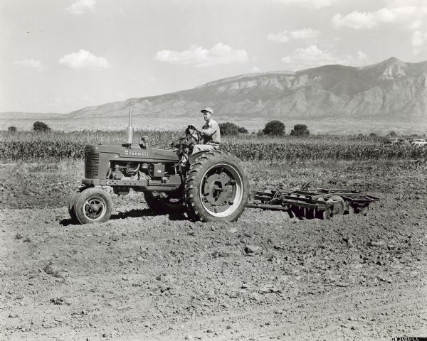 Left side profile view of a man driving a Farmall H tractor pulling a 24 6-foot, wheel-controlled offset disk harrow for a U.S. Soil Conservation Albuquerque demonstration. In the background cars are parked on the right near the cornfield. Mountains are in the far background.