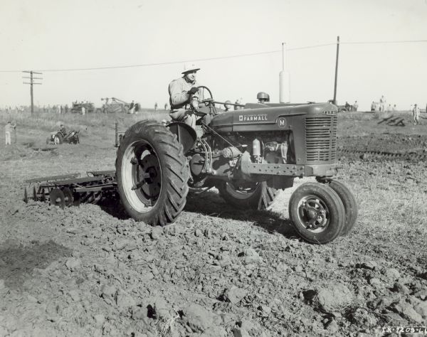 Man driving Farmall M with No. 10-A disk harrow at "Friend of the Land" meet at Oklahoma City. Other men and tractors are in the background.