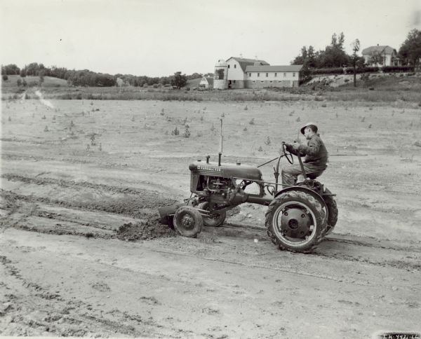 Man using Farmall Cub tractor with grading attachment in a field. Farm buildings are in the far background.