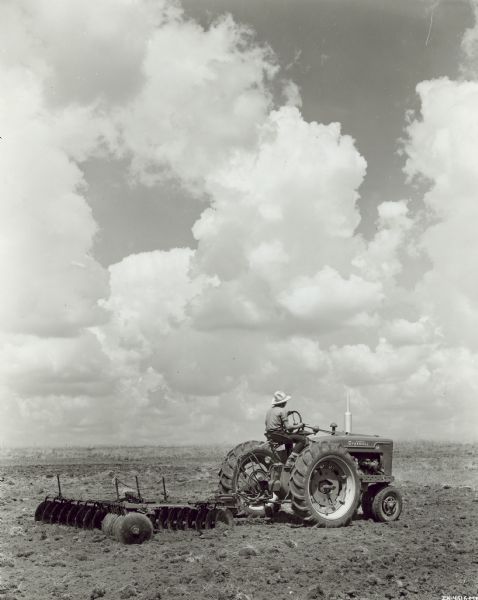 Three-quarter view of right side of Farmall M tractor with 8 foot No. 10A disk harrow equipped with remote control hydraulic cylinder. Large fluffy white clouds are in the sky.