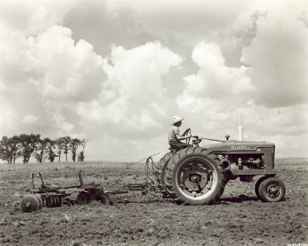 Man wearing a straw hat using a Farmall M tractor with 8 foot No. 10A disk harrow equipped with remote control hydraulic cylinder. Large fluffy white clouds are in the sky.