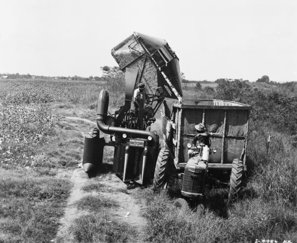 View from front of man driving a Farmall A tractor pulling a wagon. Beside the wagon on the left is a man driving a cotton picker. The cotton picker basket is tipped over and is dumping cotton into the wagon. A sign on the front of the cotton picker reads: "The Oaks Plantation — MP No. 1 — Price C. McLemore." Farm buildings are in the far background.