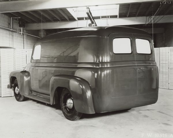Three-quarter view from left rear of Model L1-L2 cab parked indoors. Folding screens have been set up as a background in the industrial room which has exposed beams in the ceiling, and in the center, light from what may be a skylight. The "International" logo is on the hubcaps. The windows have been blocked with white paper.