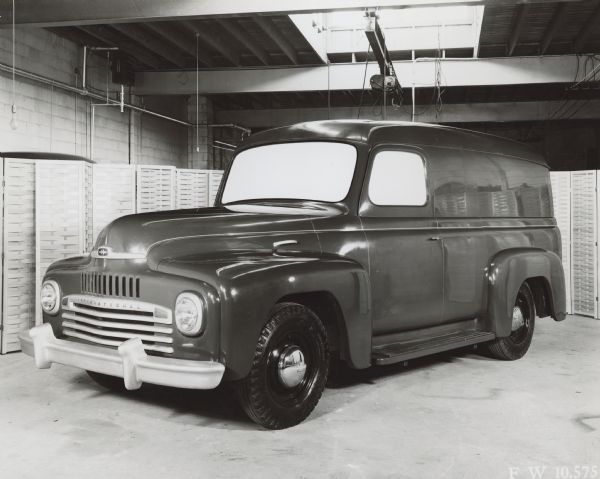 Three-quarter view from front left of a Model L1-L2 cab parked indoors. Folding screens have been set up as a background in the industrial room which has exposed beams in the ceiling, and in the center, light from what may be a skylight. The "International" logo is on the hubcaps.  The windows have been blocked with white paper.