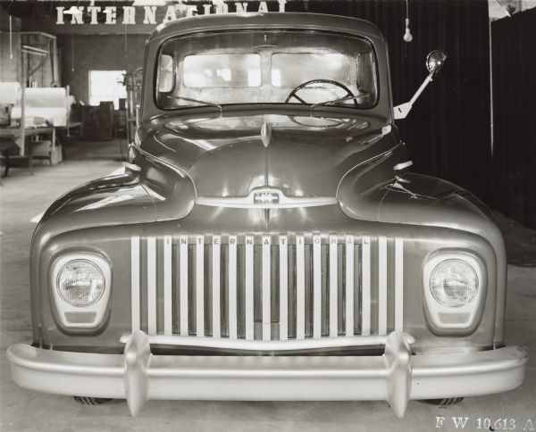 Front view of the new design cab for post WWII trucks. Dark curtains are in the background along the right for a backdrop. The "International" logo is on the front grill. In the background is a large sign for "International" hanging high on the wall in an industrial area of the factory.