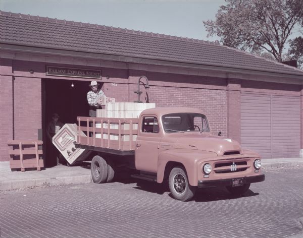 Three-quarter view from front right of a man loading or unloading a L-120 STD model truck with stake body. The truck is backed into the open door of a brick building. The sign above the doors reads: "Railway Express Agency."
