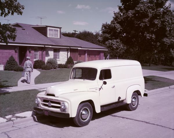 Slightly elevated view from street of an R-120 truck with panel body parked at the curb. A man is walking from the truck down the front walk of a house. He is wearing a uniform and carrying packages.