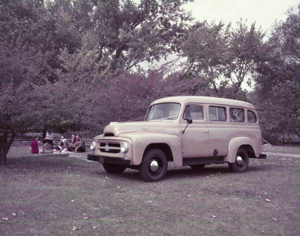 Three-quarter view from front left of an International R-110 Station Wagon parked on a lawn. In the background is a group of people enjoying a picnic.